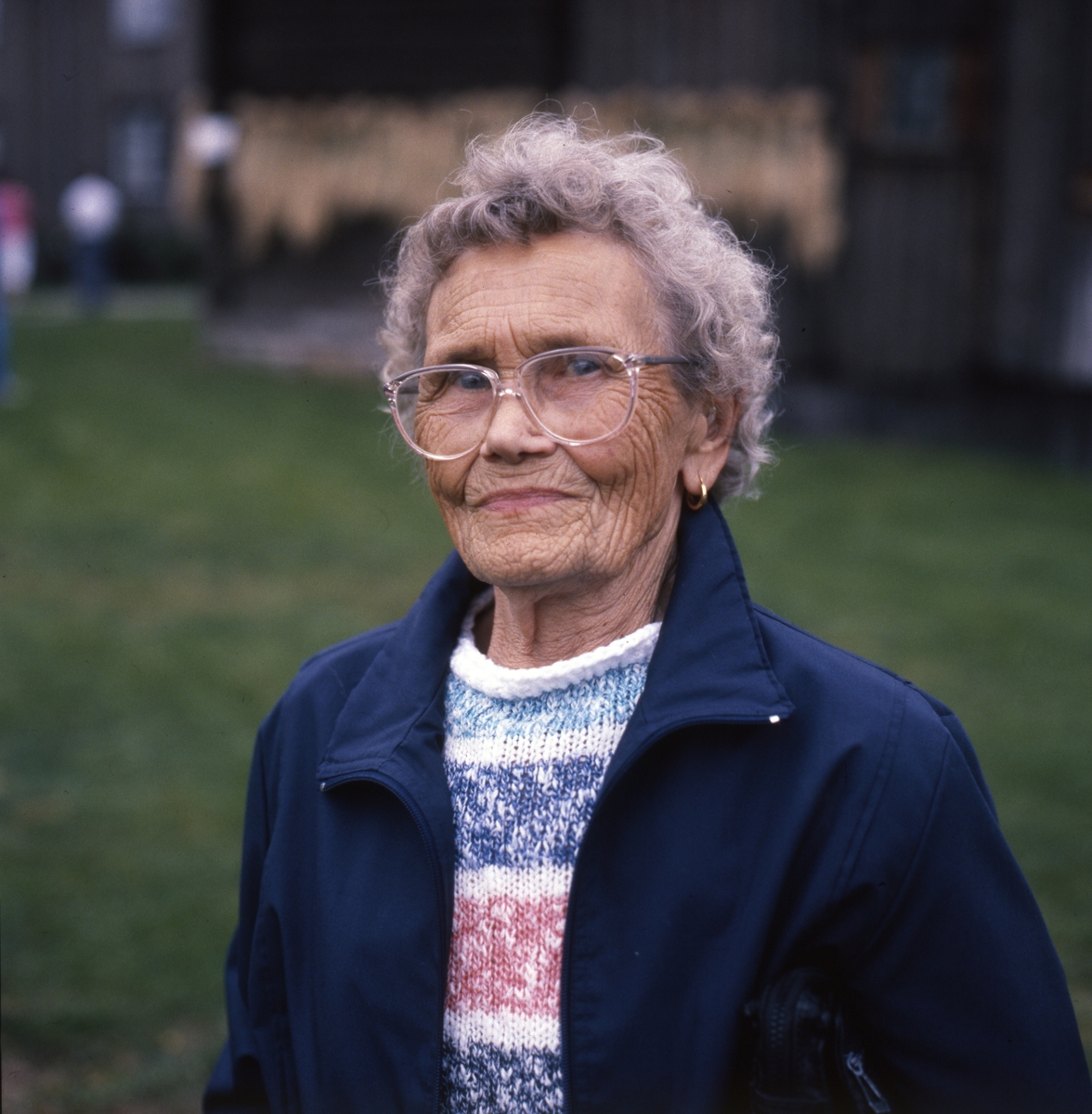 Porträtt av Brita 90 år, 1989. En kvinna med kortklippt, permanentat hår och glasögon. Hon är iklädd en stickad jumper och fritidsjacka.