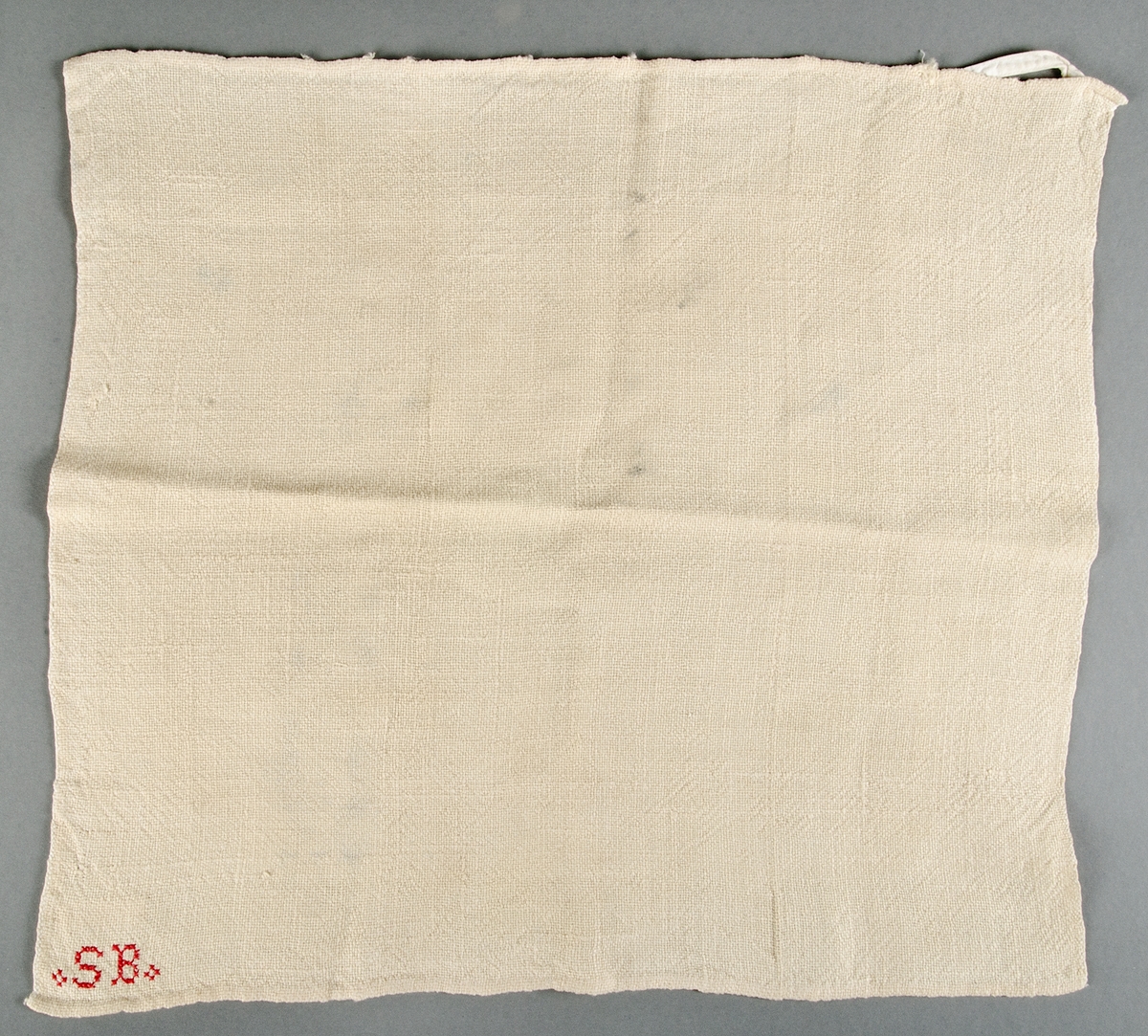 Säckhandduk av tuskaftat gulvitt linne. Upptill i ena hörnet är band till hängare fastsytt. Nertill i ena hörnet rött korsstygnsbroderi SB. Fläckar.