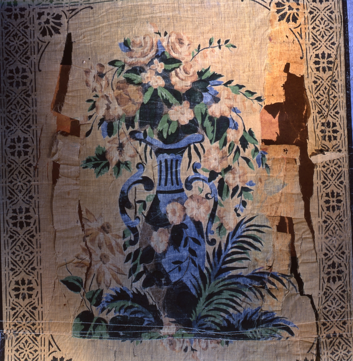 Del av rullgardin med schablontryckt mönster i form av en stor blå vas med blommor, mot ljus botten.