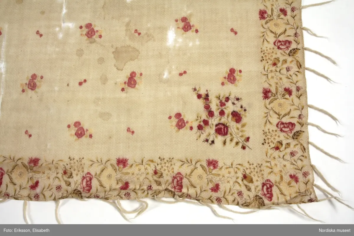 Kvadratiskt halskläde eller mindre schal av vit tuskaftad yllemuslin med svagt tryckt bikakemönster över hela  bottentyget.  I mittspegels strödda  små blommor i rosa och gulgrönt. I bården sirlig blomslinga med rosor och nejlikor i rosa och gulgrönt med tunna bruna stjälkar.  Runtom gles iknuten frans av vitt ullgarn.
Anm. Bristningar på flera ställen, bl.a en lång reva.

De tunna fina muslinschalarna  med tryckt mönster kom in i det folkliga modet som brudschalar vid mitten av 1800-talet.  En del var importerade men tillverkning fanns också i Göteborg.
 Tryckaren J.J.Amelung i Stockholm var den första som kunde trycka på tunt ylletyg 1834. Han gjorde dukar med mönster efter de sk. kashmirschalarna. Se Henschen: Kattuntryck , 1992 sid 70-71.
/Berit Eldvik 2012-01-05