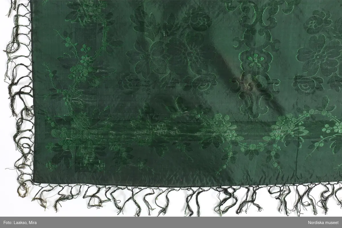 Kvadratiskt halskläde i grönsvart sidentaft med jacquardvävt mönster med inslagsflotteringar  i grönt, spegeln indelad i 4 stora rutor med ornamenterade bårder och en stor blombukett i varje ruta. Runt kanten blomslinga. Tvåknuten frans av tvinnat silke i 2 gröna nyanser.
Likadant halskläde i andra färger se 148 963 från Ilsbo, som är hallstämplat i Stockholm
/Berit Eldvik 2011-11-10