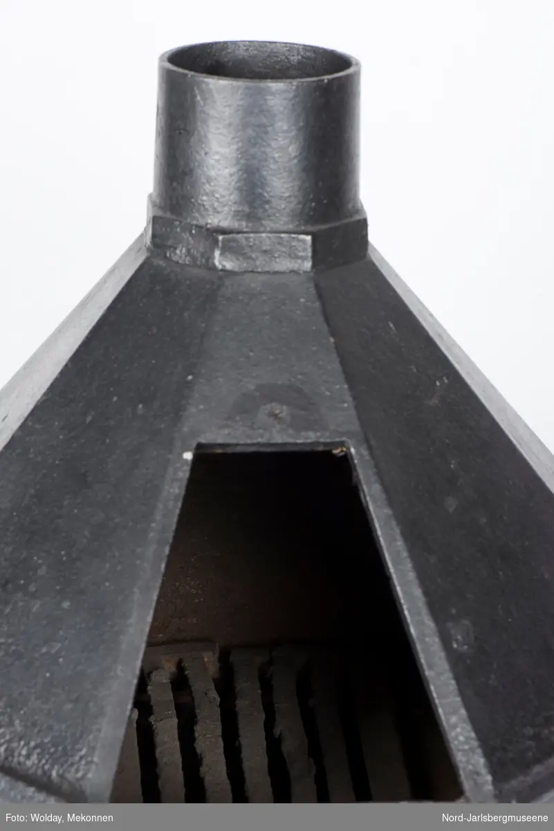En liten, åttekantet ovn til å varme strykejern. En av sidene er dør til obvnskammeret, røyktut midt opp.  Askeskuff.