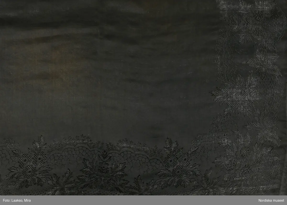 Kvadratiskt halskläde av svart tunn sidensatin med slät omönstrad mittspegel och jacquardvävt blommönstrad bård,  med iknuten frans av svart tvinnat silke. 
/Berit Eldvik 2011-11-30