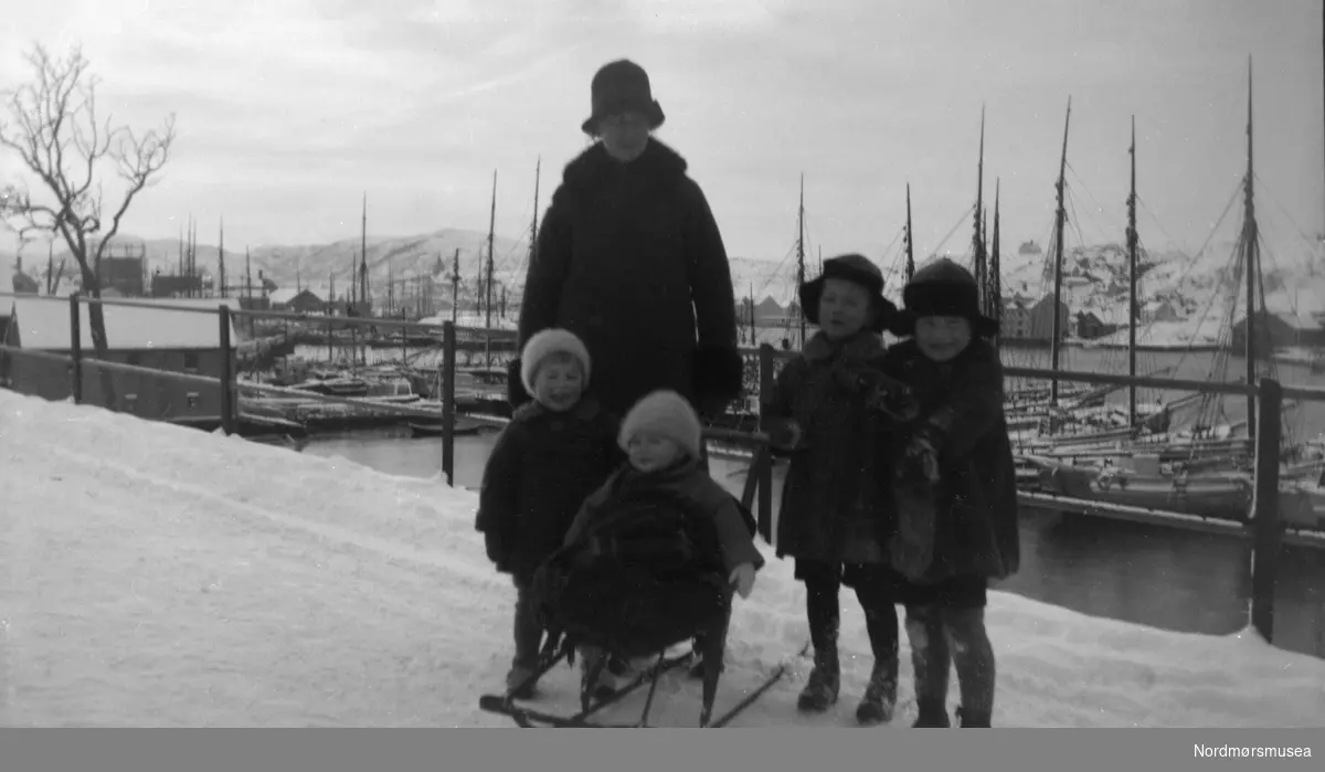 Foto av en kvinne ute med fire små barn med spark. Datering er usikker men trolig rundt 1917-1920. Bildet ser ut til å være fotografert langs Kranaveien i Vågen på Kirkelandet i Kristiansund. Fra Nordmøre museums fotosamlinger.
