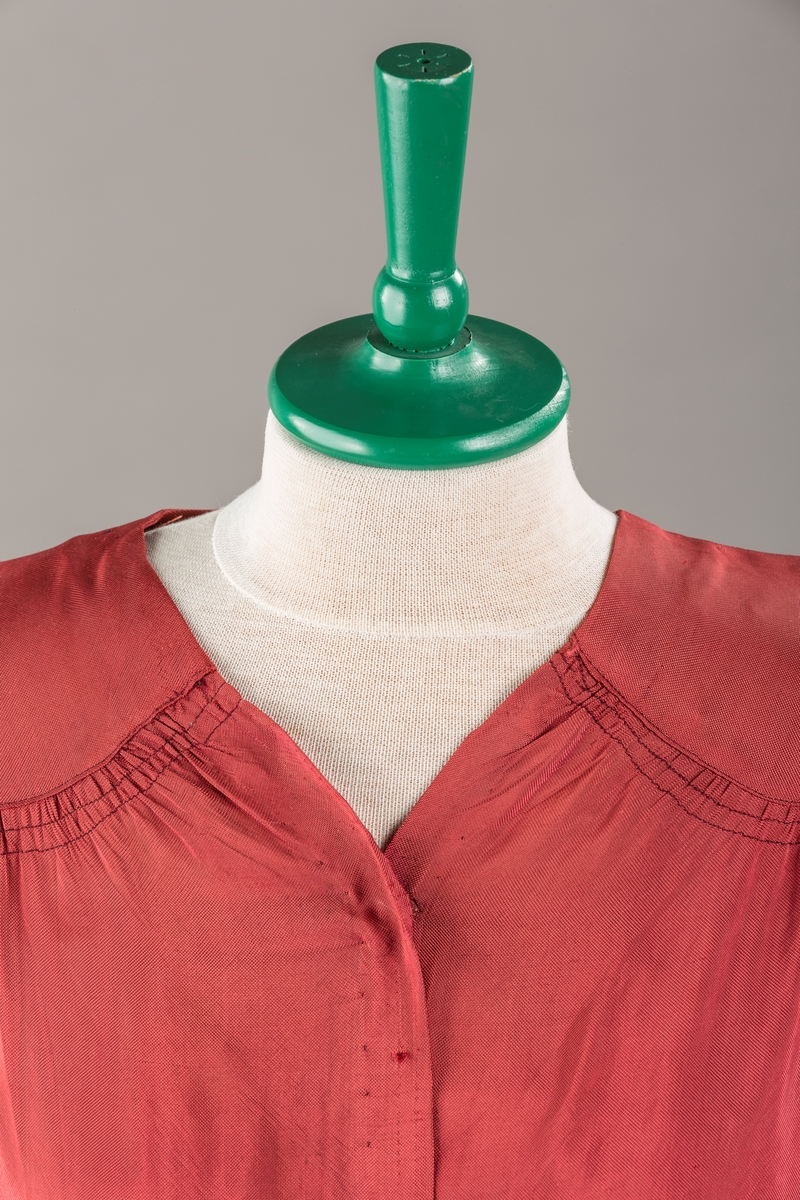 Form: Glatt, klokkeformet skjørt. Forstykkene rynket til buede bærestykker foran. Overdel rynket til skjørt. Korte ermer. Lukket med trykknapper. Belte fastsydd.