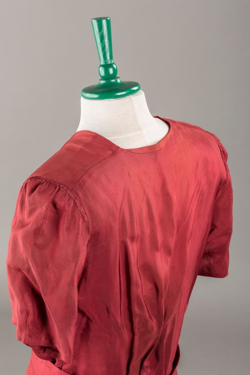 Form: Glatt, klokkeformet skjørt. Forstykkene rynket til buede bærestykker foran. Overdel rynket til skjørt. Korte ermer. Lukket med trykknapper. Belte fastsydd.