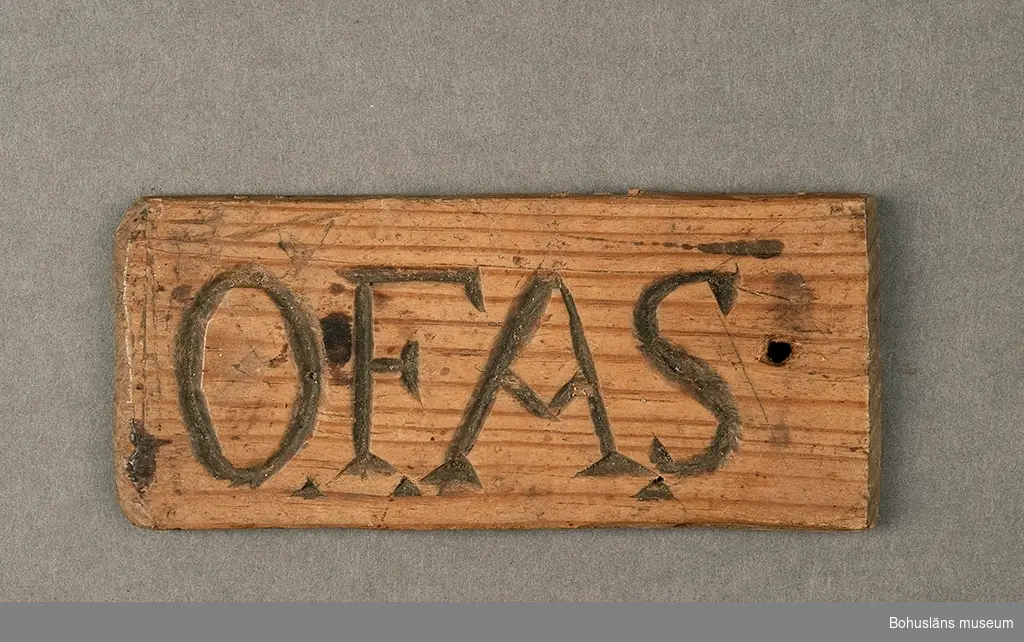 Bricka med initialerna O.F.A.S (i djuprelief) att ha tillsammans med nycklar. På brickans baksida mönster av olika karvsnitt. Eventuellt en träbit som återbrukats som nyckelbricka.