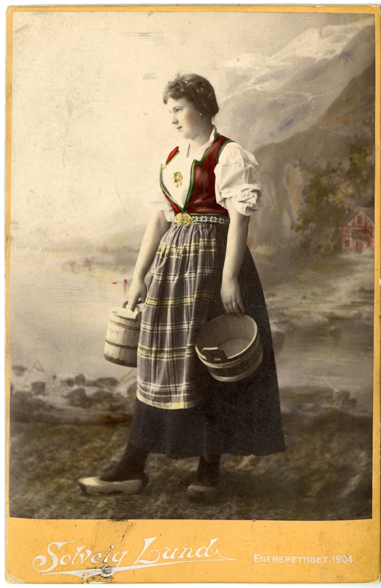Kolorert studiofotografi av kvinne med Hardangerdrakt, bærende på to tiner.