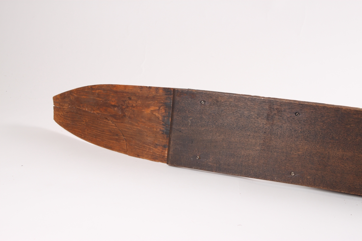 En ski med en lett krummet form og spiss i begge ender. Omtrent midt på skien er det en opphøyning i treet hvor det går et horisontalt hull gjennom skien til å feste bindingen i. På hver side av fotstegplaten og bindingsfestet går det en bred kant. Oversiden av skien er flat, med en noe buet overflate på den ene siden av skien. På undersiden av skien er det påmontert en treplate formet etter skien. Platen er festet med skruer. Skien har to grunne styrerender.