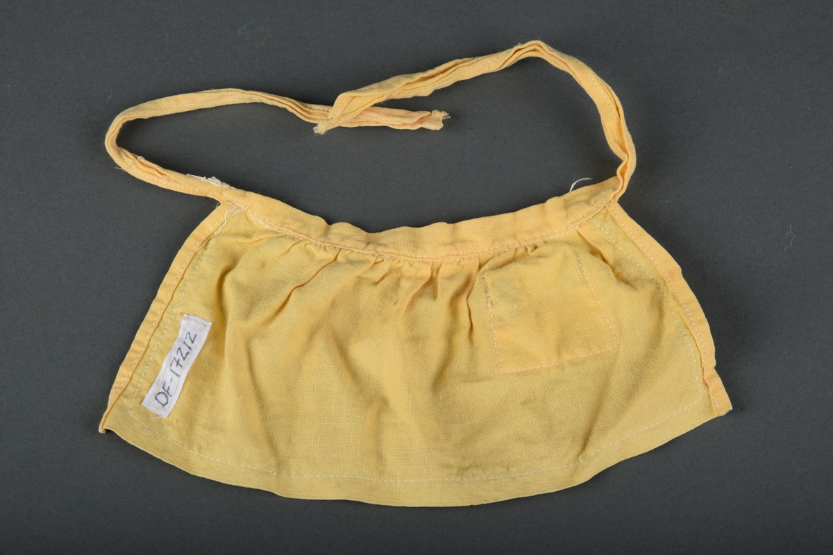 Forkle til dukke i gul tekstil. Liten lomme foran på forkleet og stropper til å knytte forkleet med. Påsydd grønt bånd langs kanten, og som detalje på lommen.