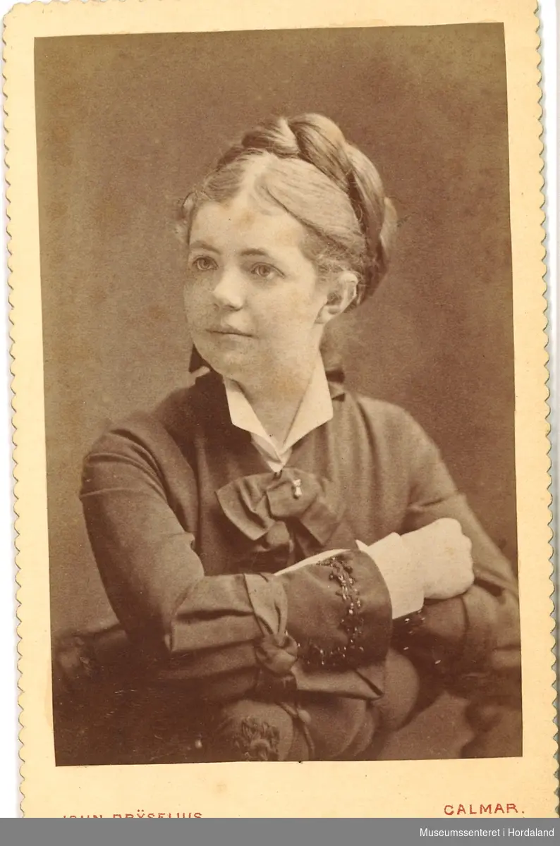 halvfigurfotografi av ung kvinne med ljos hårflette rulla opp, mørk drakt med kvit krage og sløyfe, armane i kross