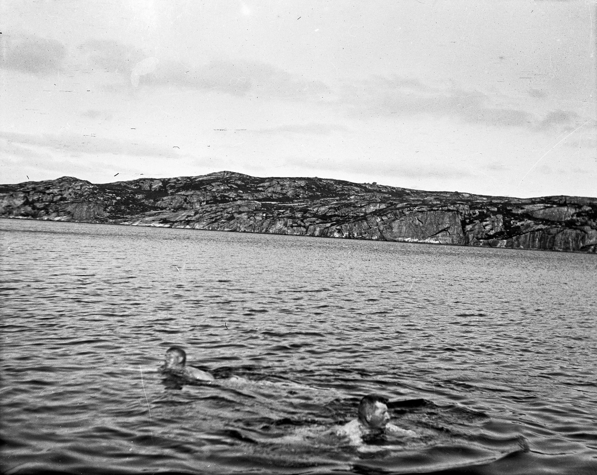 Kystlandskap. 2 menn bader i sjøen i forgrunnen. Landskap i bakgrunnen.