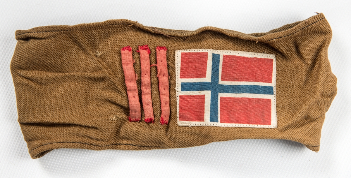 Armbind i brunt bomullstøy. Påsydd tre tynne raude striper og det norske flagget. Skreve med penn OSP. Var sett på ermet til VFF 09535 Uniformsjakke (med sikkerheitsnål).