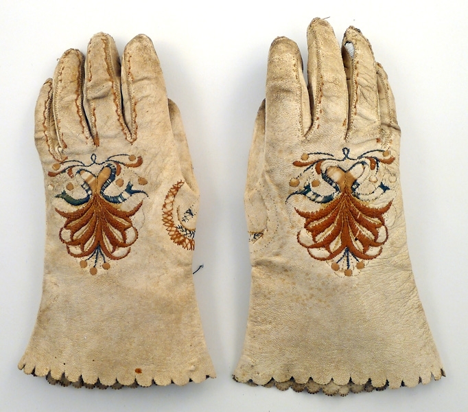Handskar av vitt skinn med broderat mönster i grönt och två nyanser brunt. Även brun rand runt fingersömmarna. Broderiet delvis bortslitet. Smutsiga
