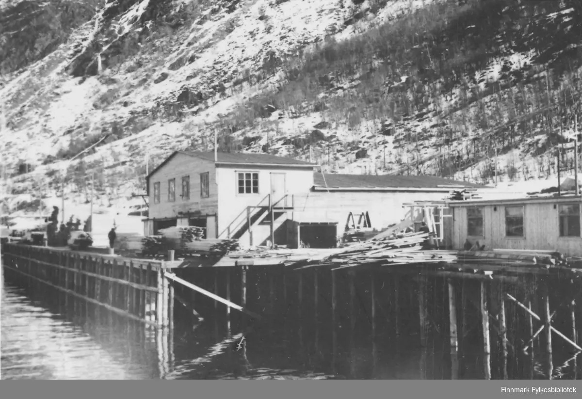 Før og etter 1944, Nordland, Troms. Bildet er tatt fra sjøen og inn mot en kai. Bildet viser noen trebygninger som oppgis å være henholdsvis et høvleri, spisebrakker,kontorer. Trematerialer ligger stablet rundt bygningene. Snøen ligger ennå i åsen bakenfor kaia.