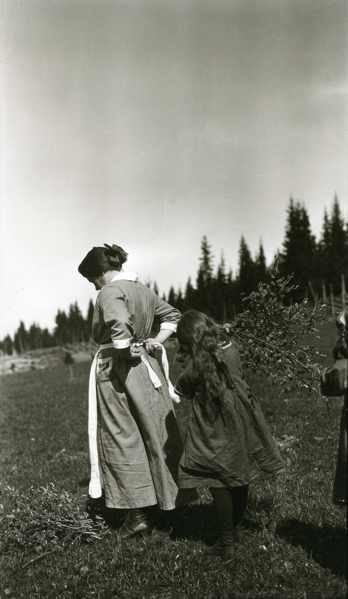 Kvinne og jentunge i Galåsen. Begge står med ryggen til. Kvinnen, Oline Galaasen (1872 - 1944) knyter på seg forkleet. Jenta, Ingebjørg Buflod f. Galaasen (1915) el. Målfrid Hauge f. Galaasen (1914) (sannsynligvis Målfrid) står med kvister i handa.