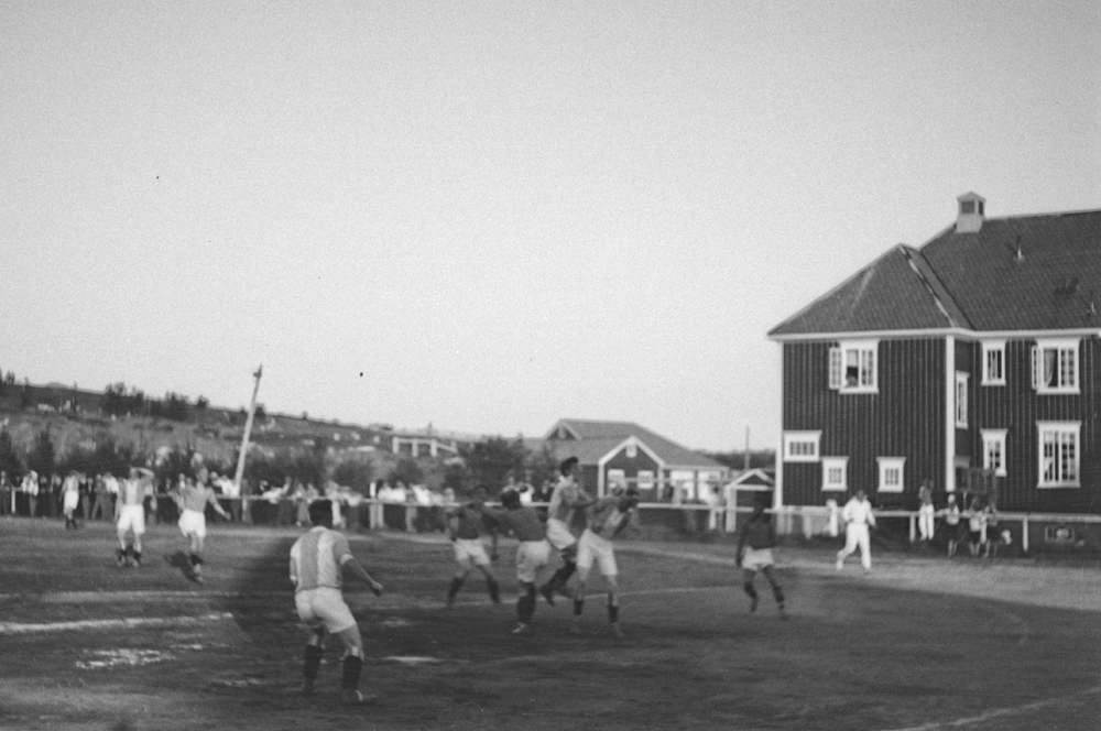 Fotballkamp. Dette er en kamp i 2. runde i NNM i fotball mellom Ski og fotballklubben Glimt Bodø og Mosjøen IL på Skøytebanen idrettsplass ved sykehuset i Bodø. Vi ser Rensåsen i bakgrunnen. Resultatet ble 4-2 til Glimt. Kampen ble spillt søndag 5. august i 1934.  Ref: Nordlandsposten 6. august.