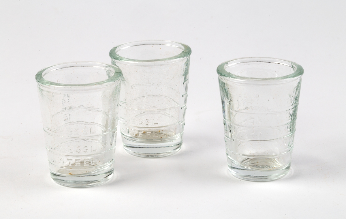 Eske av kartong med lokk. Inndelt i 24 rom med 23 måleglass. Hvert måleglass med innstøpt gradering på ene siden opp til 20 ml, på annen side 1 TE EL / 1/2 E SSL / 1 ES SL.
Esken er holdt sammen med hyssing. På lokket notert med blyant: 5704