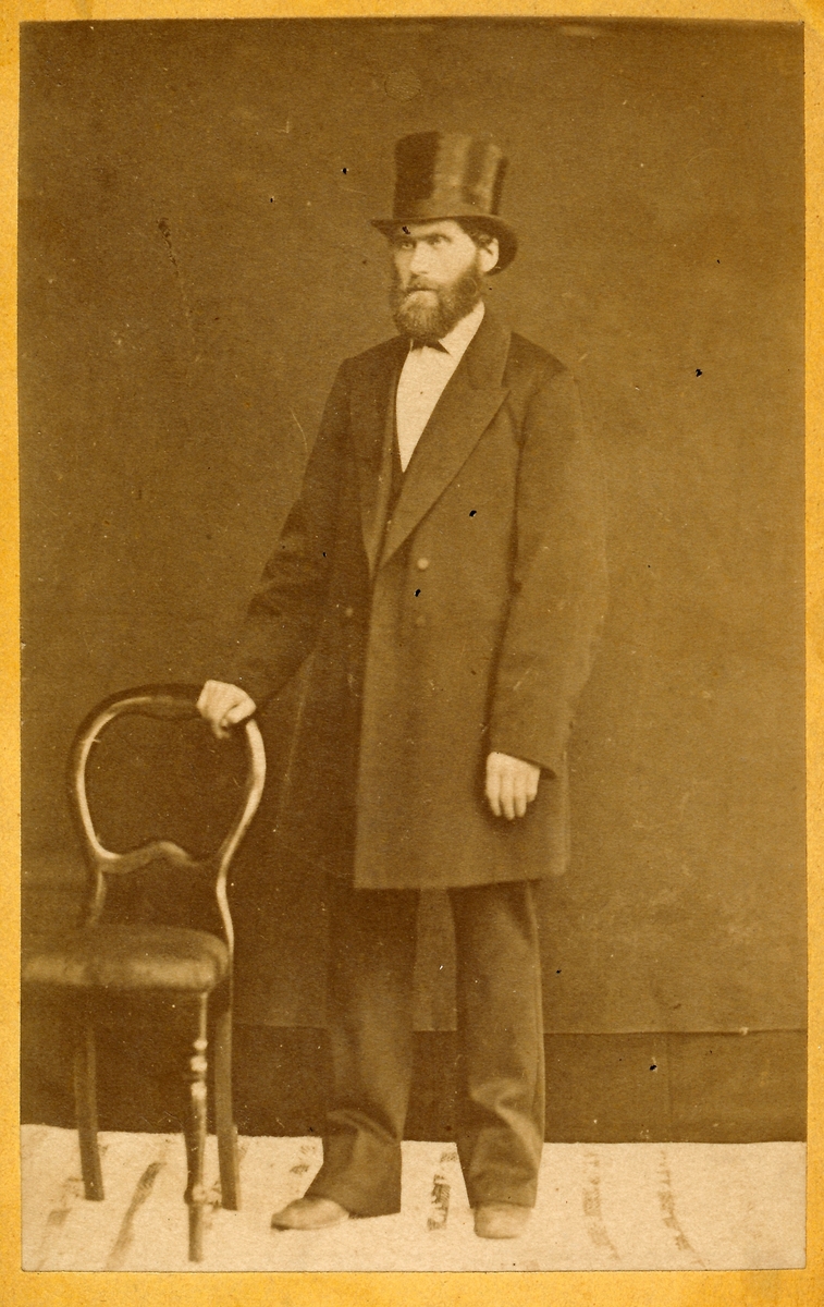 Portrett, Visittkort. Mann med frakk, skjegg og flosshatt står ved siden av stol. Baker Peder Pedersen (f. 1841) på Mo.