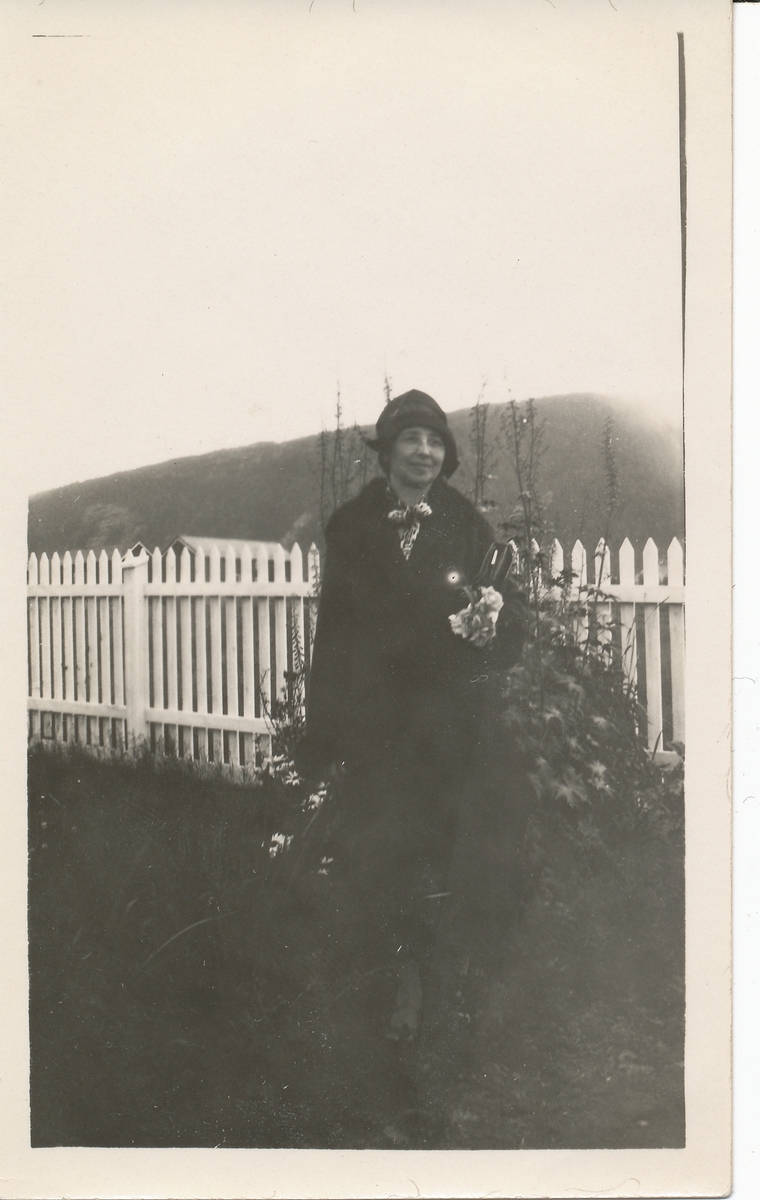 Kvinne i hatt og kåpe står ved et stakittgjerde i hage. Frk Leinum.