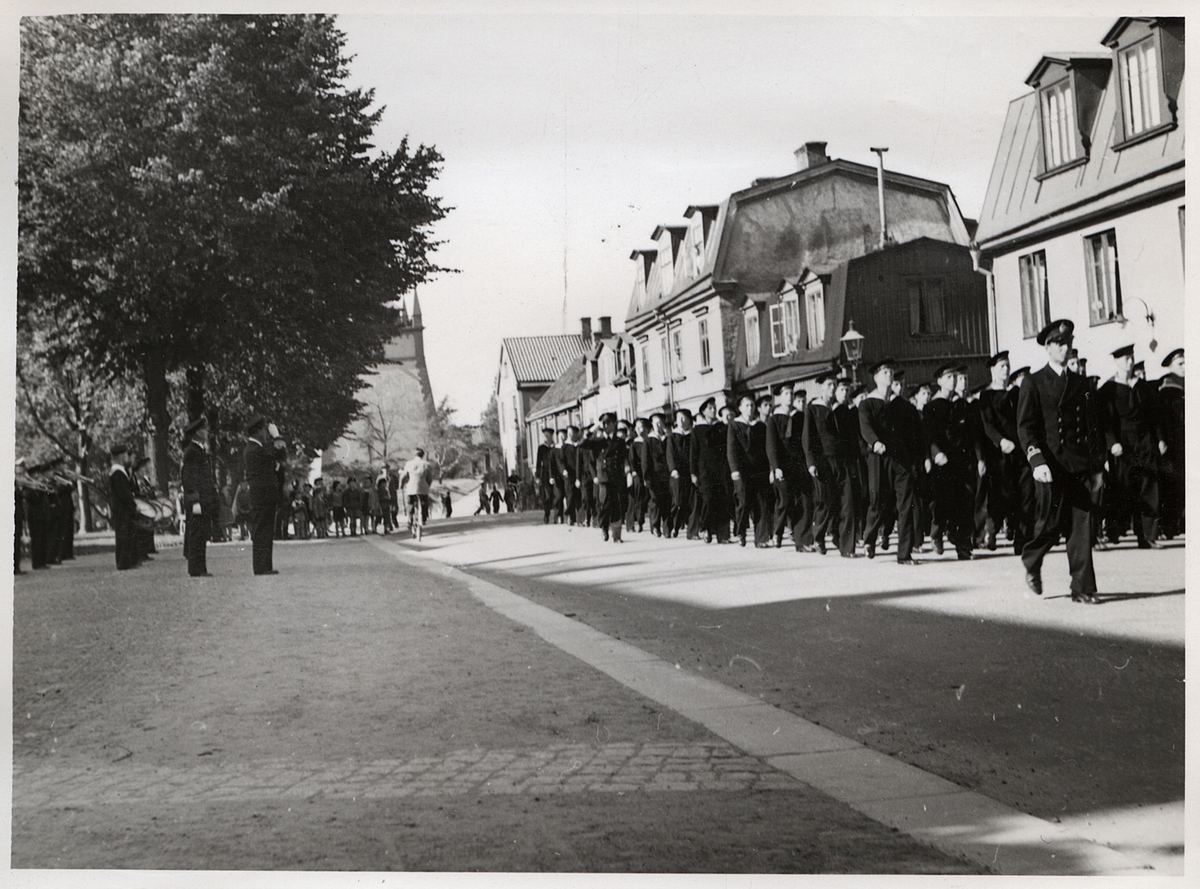 Bilden visar en militärparad som marscherar i stadsmiljö, närmare bestämt på Vallgatan i Karlskrona. Ledet med unga rekryter ses till höger i bild och sträcker sig från förgrunden in i bakgrunden. I förgunden ses också ett högre befäl, en löjtnant, som går med i marschen. Längre bak i ledet finns ytterligare ett befäl som går med och han hälsar med en honnör till sina kollegor som står i mellangrunden till vänster i bild. Dessa hälsar på samma sätt. Här syns också medlemmar ur marinens musikkår med pukor och blåsinstrument. 
I bildens bakgrund ses civilbefolkning,bland annat barn som tittar på paraden och en man på cykel. Bakom ses träd som delvis skymmer Amiralitetsklockstapeln i Amiralitetsparken.