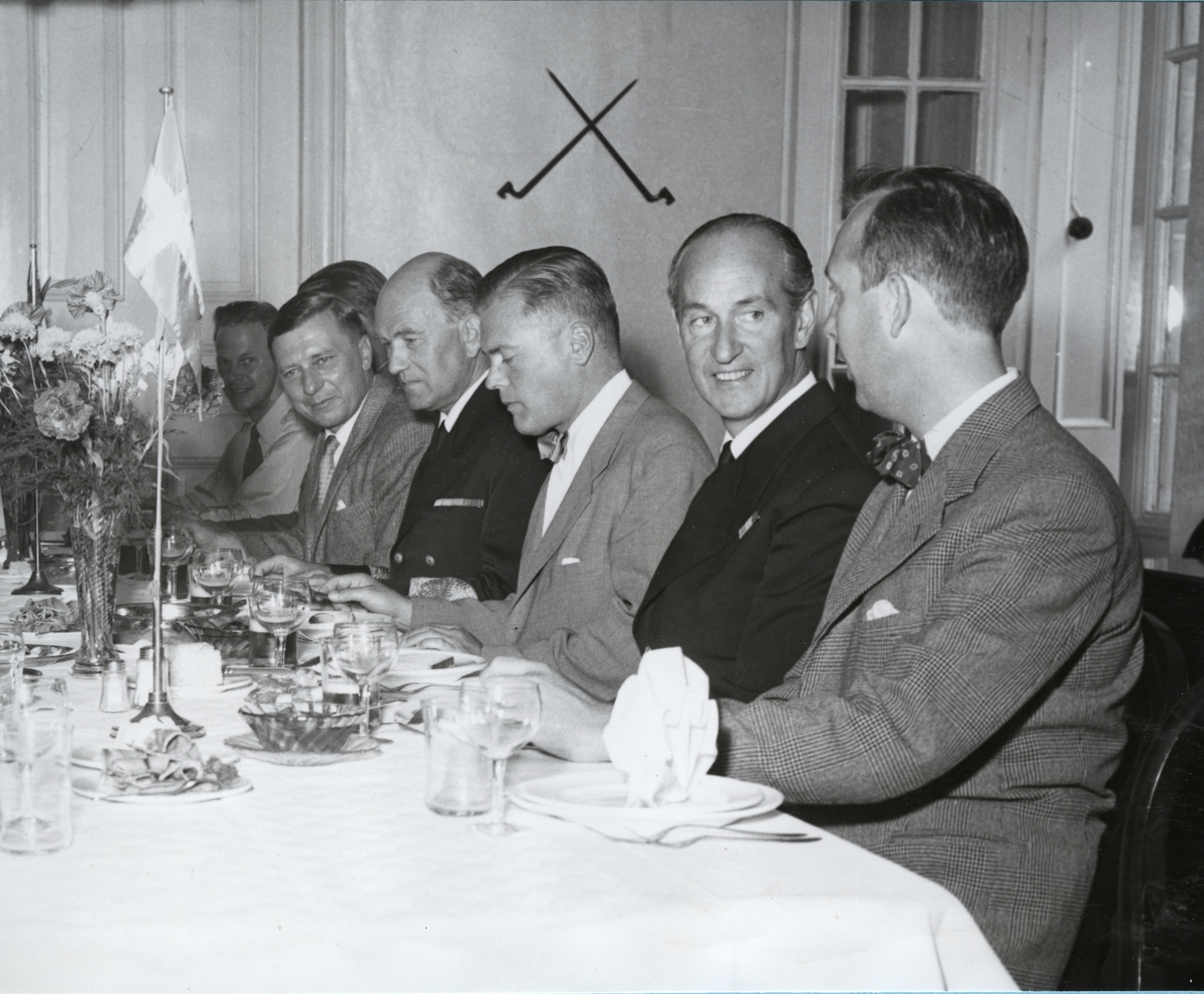 En rad av män sitter ned vid ett dukat middagsbord. Kommendörkapten Lind af Hageby (andra från höger) och konteramiral Berthelsson (i mitten) klädda i uniform.