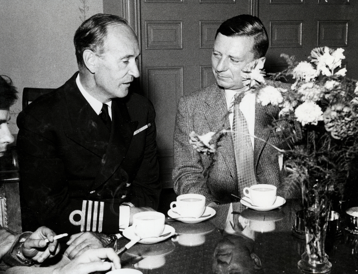 Kommendörkapten Lind af Hageby sitter vid ett kaffebord och konverserar med en kostymklädd man. Tre kaffekoppar syns på det runda bordet tillsammans med en vas med blommor.