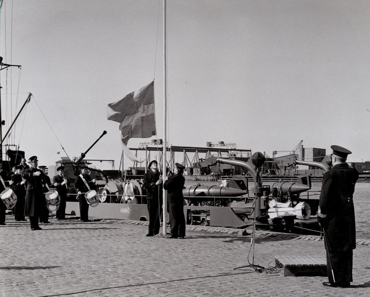 Två flottister halar eller hissar en svensk flagga. Bakom dem spelar en musikkår och framför dem står en konteramiral och bevittnar händelsen. Samtliga är klädda i uniform. Längs med kajen ligger minsveparen HMS Hanö (M51) förtöjd.