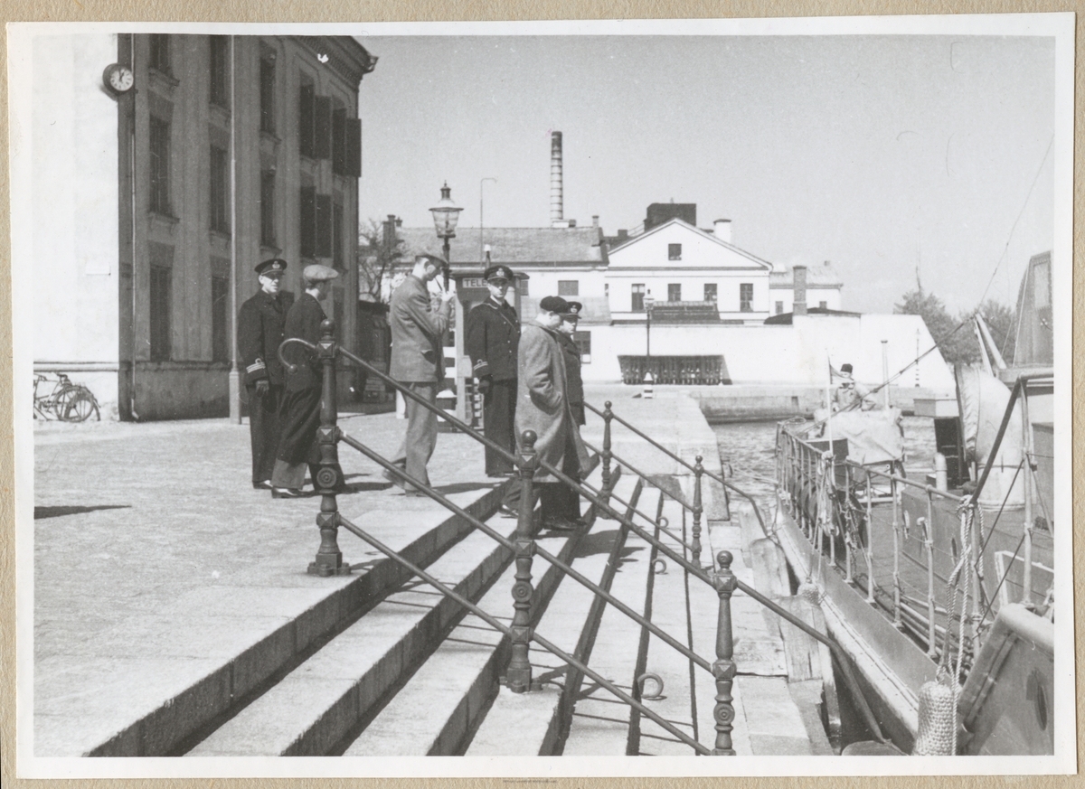 Civila och militära män, bland annat en kapten och en löjtnant, på väg ned för trappan vid kungsbron. Vid trappan ligger ett örlogsfartyg förtöjt. I bakgrunden syns Hollströmska magasinet och lite längre bort byggnader tillhörande bataljon Sparre, t.ex. matinrättningen.
