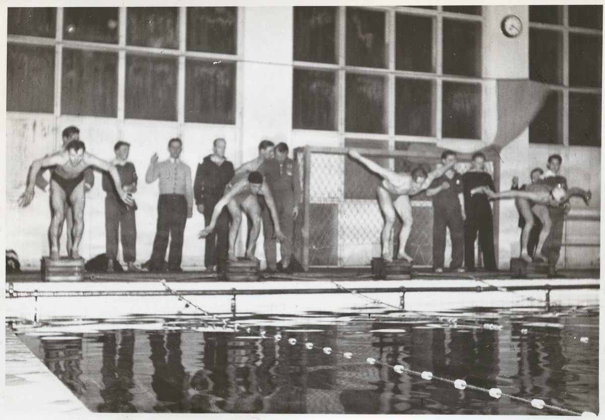 Fyra män i badkläder i färd med att hoppa i en simbassäng under en simtävling. Bakom dem står flera män och tittar på.