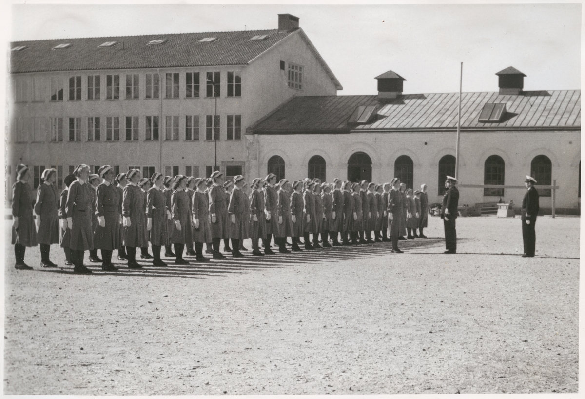 Uniformsklädda lottor står uppställda på bataljon Sparres kaserngård. Framför dem står två militärer av högre rang, även dem i uniform. I bakgrunden syns exercishuset tillhörande bataljon Sparre, med putsad fasad och rundbågiga fönsteröppningar.