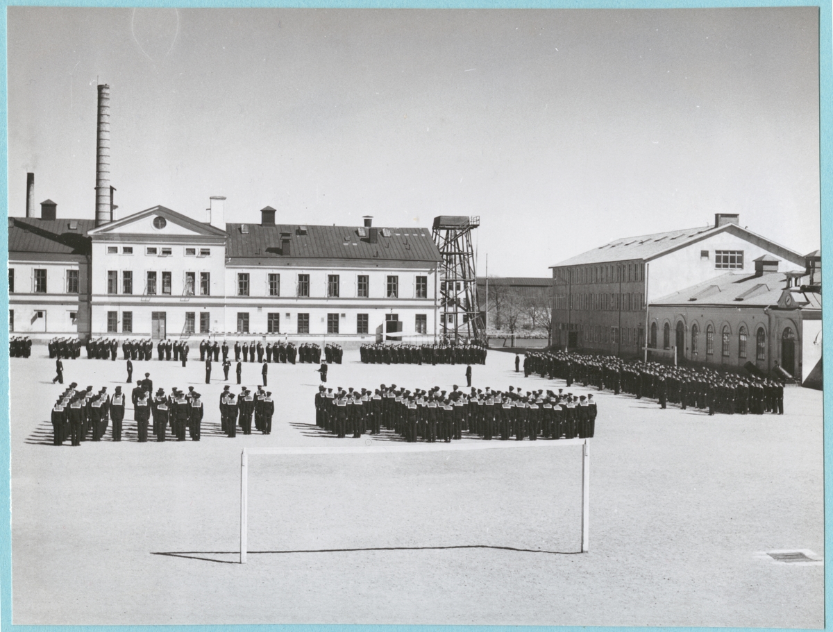 Uniformsklädda flottister står uppställda på bataljon Sparres kaserngård. Kaserngården omgärdas av matinrättningen och exercishuset.