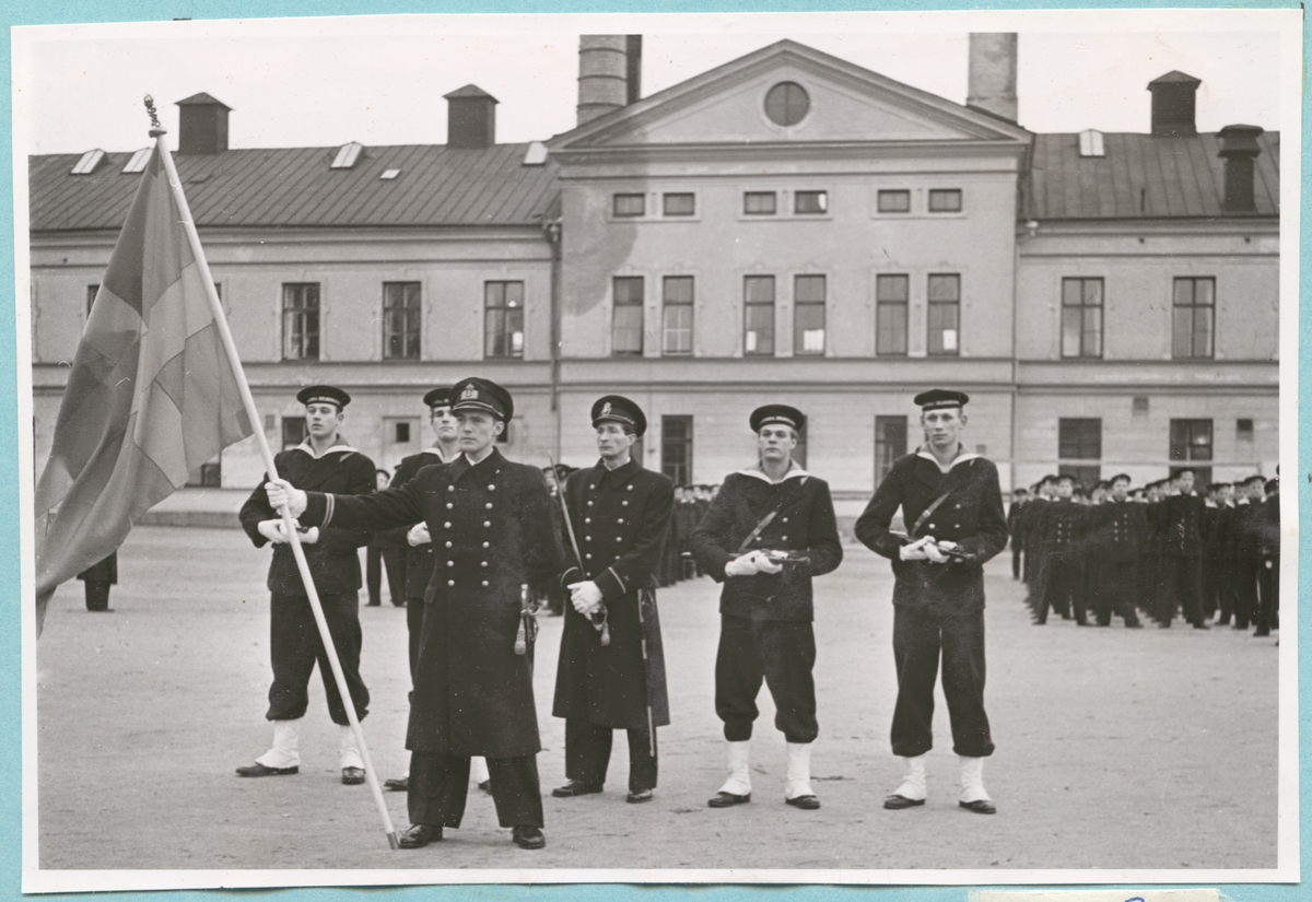 Fem uniformsklädda och beväpnade militärer står på kaserngården framför matinrättningen tillhörande bataljon Sparre. Framför de fem militärerna står ytterligare en militär som håller i en flagga. På kaserngården syns även flera uppställda militärer.
