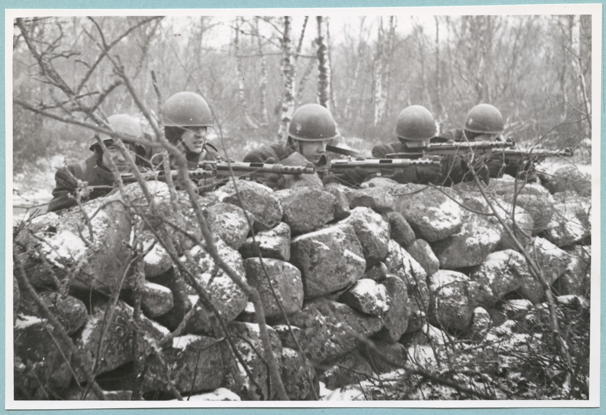 Fem uniformsklädda militärer sitter bakom en stenmur och siktar med skjutvapen under en övning. Samtliga har hjälm på huvudet. Ett tunt lager snö har lagt sig på muren.