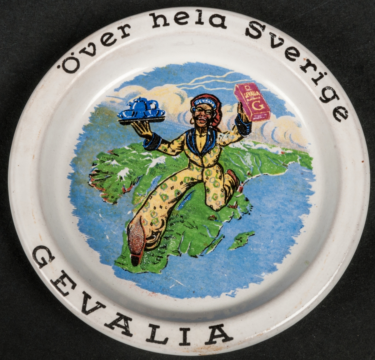 Askfat av flintgods försett med reklamtryck: "Över hela Sverige Gevalia". Bilden föreställer en mörk man som håller ett paket kaffe i handen. Förmodligen tillverkat vid Gefle Porslin i Gävle. Ostämplat.