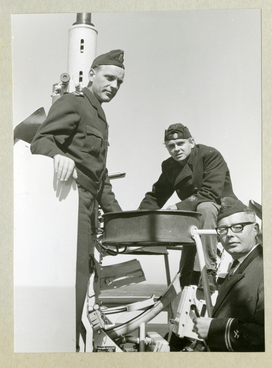 Bilden föreställer tre uniformsklädda besättningsmedlemmar vid en luftvärnskanon ombord på minfartyget Älvsnabben under långresan 1966-1967. Nederst till höger syns flaggartillerimästaren Evert Ahlin, till vänster en högbåtsman och i mitten en menig artillerihantverkare.