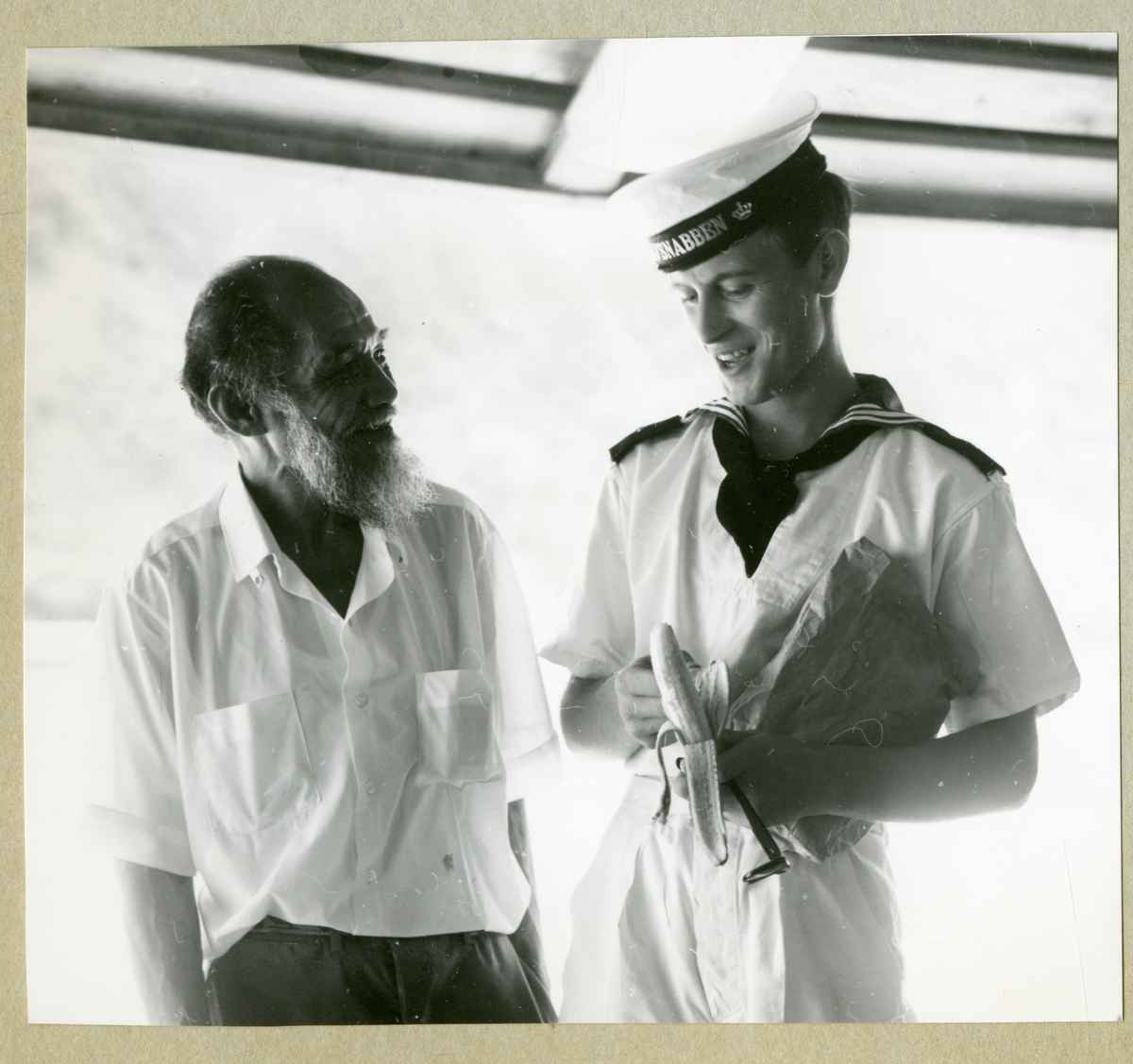 Bilden föreställer en besättningman i vit uniform som skalar en banan. Bredvid står en äldre man. Bilden är tagen under minfartyget Älvsnabbens långresa 1966-1967.