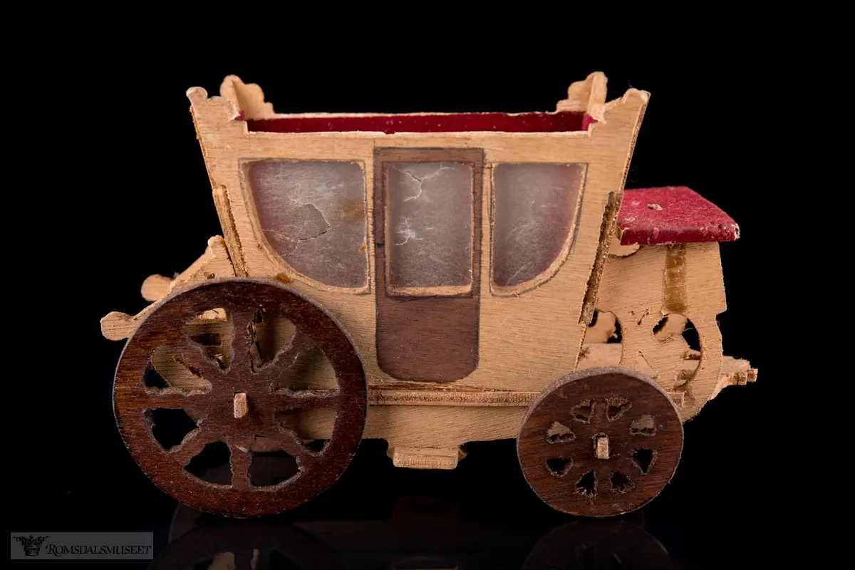 En liten leke- hestevogn i kryssfiner. To store og to små hjul, kledd med rød filt og vokspapir er brukt som vinduer.