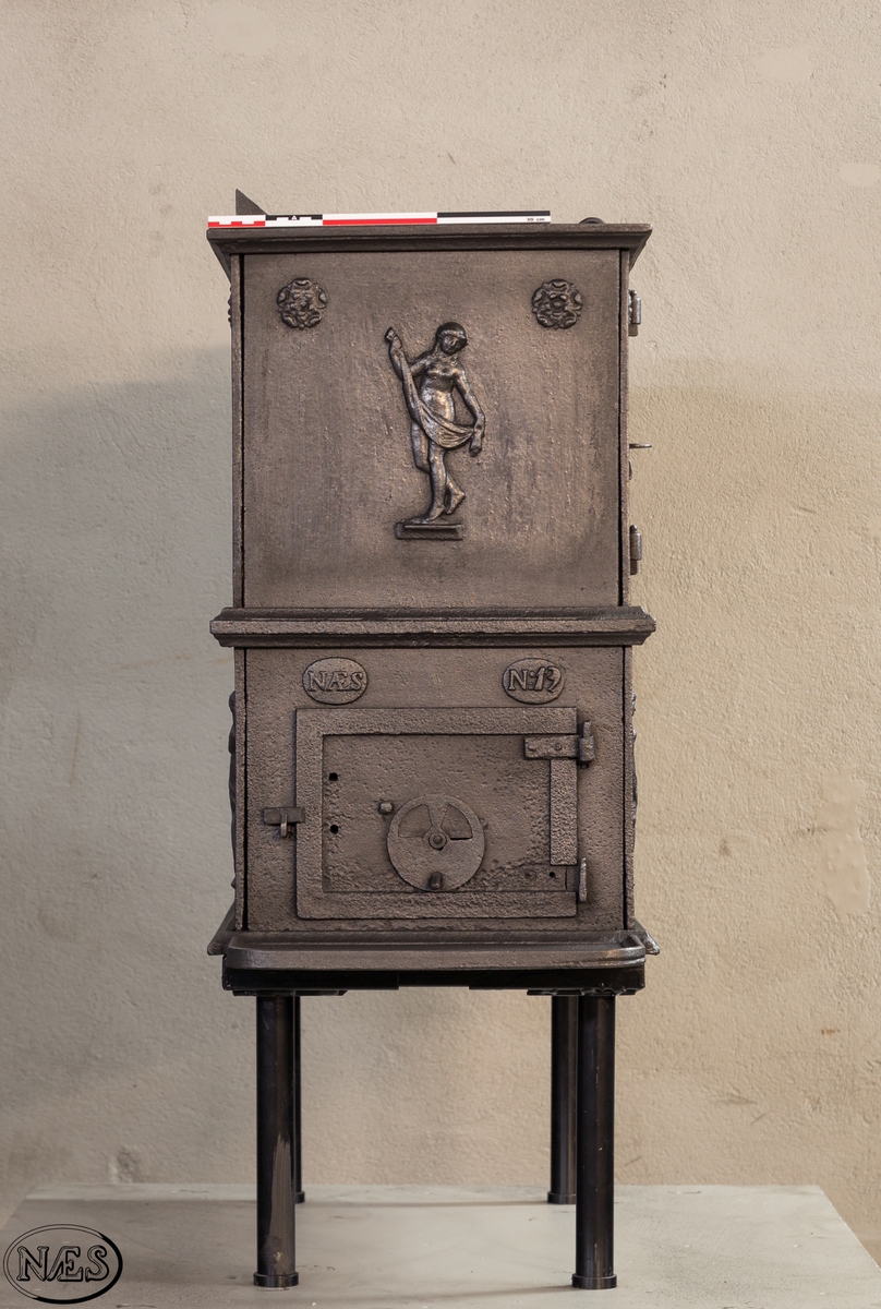 Kokeovn i to etasjer med eget fyrkammer og kokerom. Kokerommet kan åpnes med to dører på langsiden og med en luke i topplaten. Ovnsdøren til fyrkammeret er på kortsiden 1 etg. På langsidene i 1. etg er det tre muser og en engel med lette draperier på en enkel profillist. På kortsidene 2. etg er det draperikledd dansende muse på enkel profillist med en rosett i hvert øvre hjørne. Langsidene i 2.etg er dekorert med en rosett i hvert øvre hjørne.