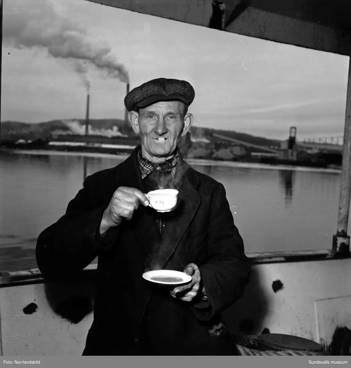 En man vid Skönviks sågverk dricker kaffe på bit. I bakgrunden syns Östrandsfabriken.