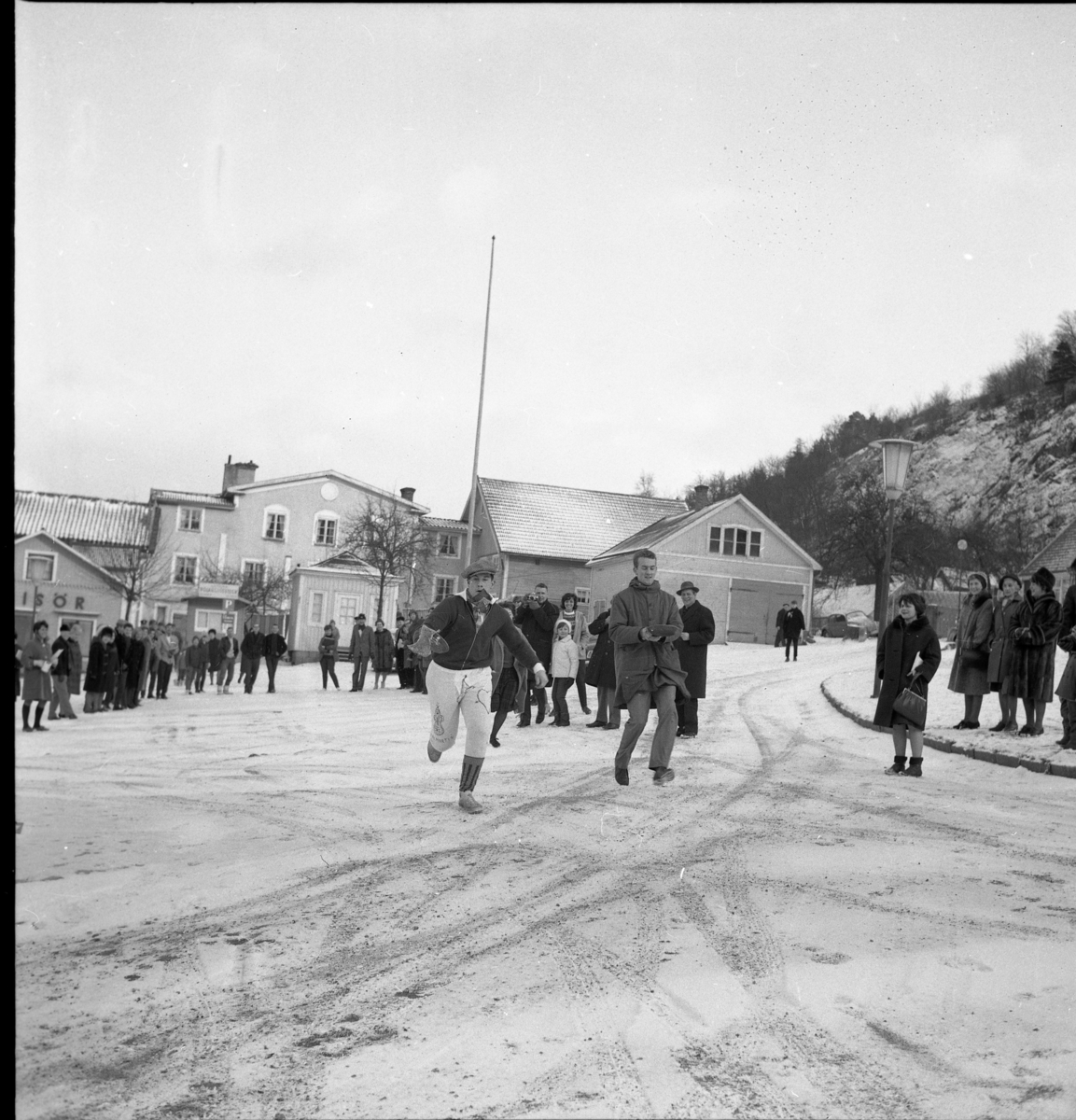 Grännaskolan arrangerar "Pannkakslopp". Två elever springer över ett snötäckt Gränna torg, från norra delen mot den södra, med en stekpanna med pannkaka i. Till vänster Johan Åkerlund och okänd till höger. I bakgrunden kvarteret Ångbåten med bl a Risbergs frisörsalong och bakom den hade Aston Bäck en cykelverkstad.