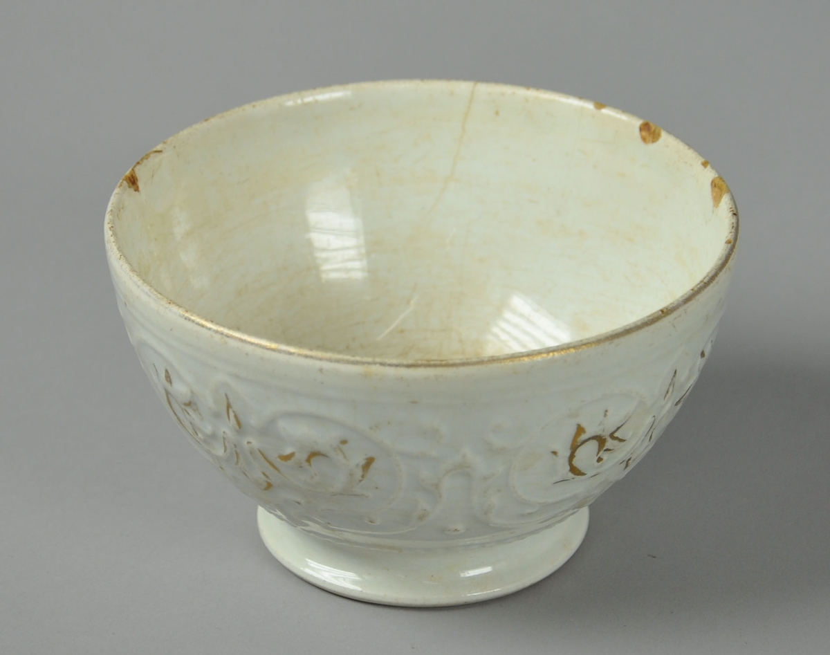 Hvit skål av glassert keramikk, med stett. Skålen har utstående dekor med spor av gullmaling.