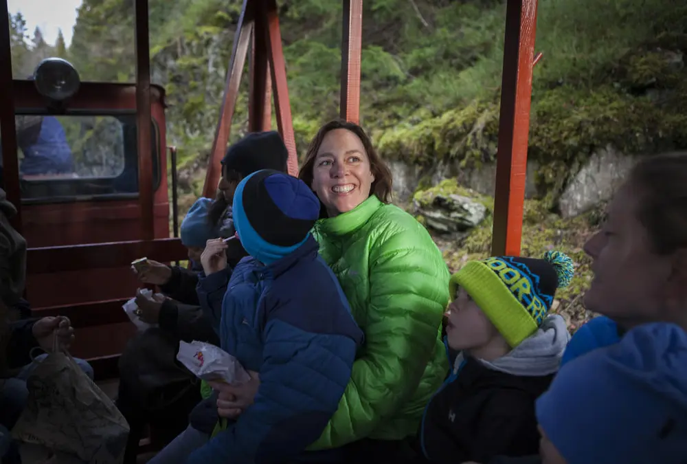 Kvinne i grønn boblejakke som smiler, og har et barn på fanget. De sitter sammen med flere i en enkel jernbanevogn.