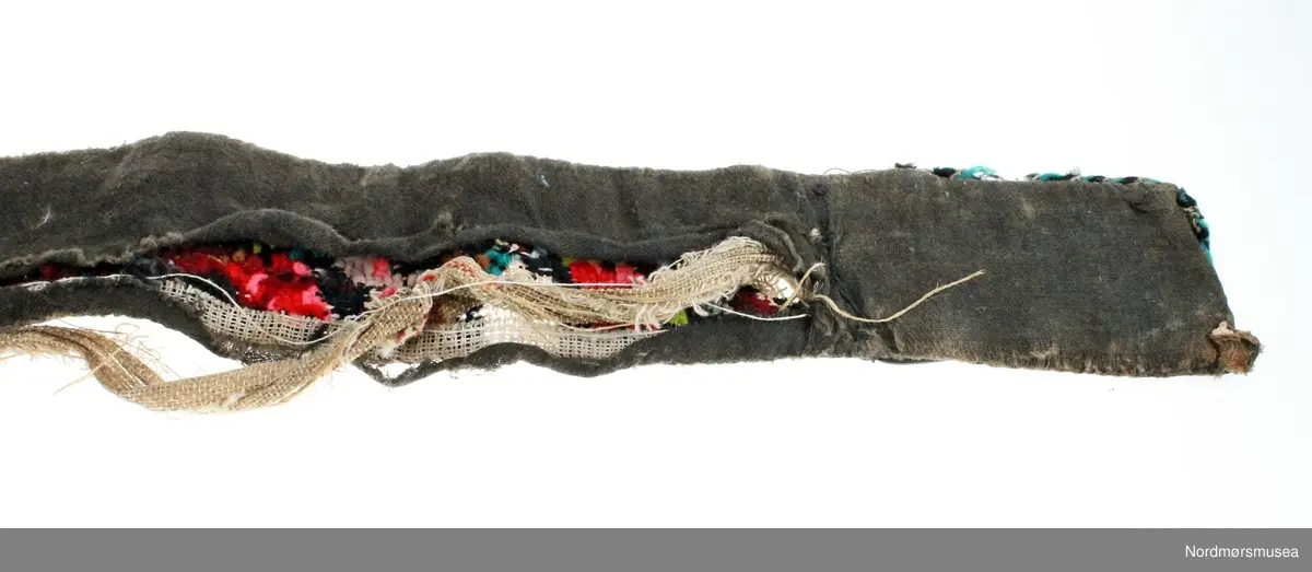 Mannsbelte brodert på stramei med blomster- og bladmotiv i mange farger. Beltet er fôret med svart ullstoff og er påsydd ei sølvfarga metallspenne. Innleggsfor i lin. Beltet er svært slitt og fôret er revnet.