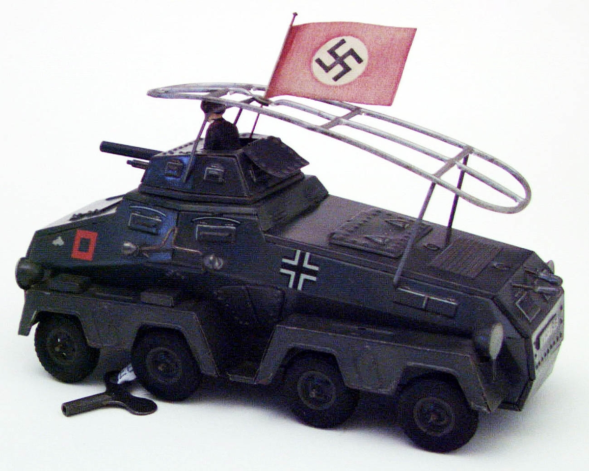 Leksaksstridsvagn av lithograferad plåt. Mörkt grön med dekorationer i rött och vit/svart. Okänd tillverkning. Saknar stämplar. Till stridsvagnen finns en soldat, förmodligen av elastolin, en nazistflagga samt en nyckel för uppskruvning.