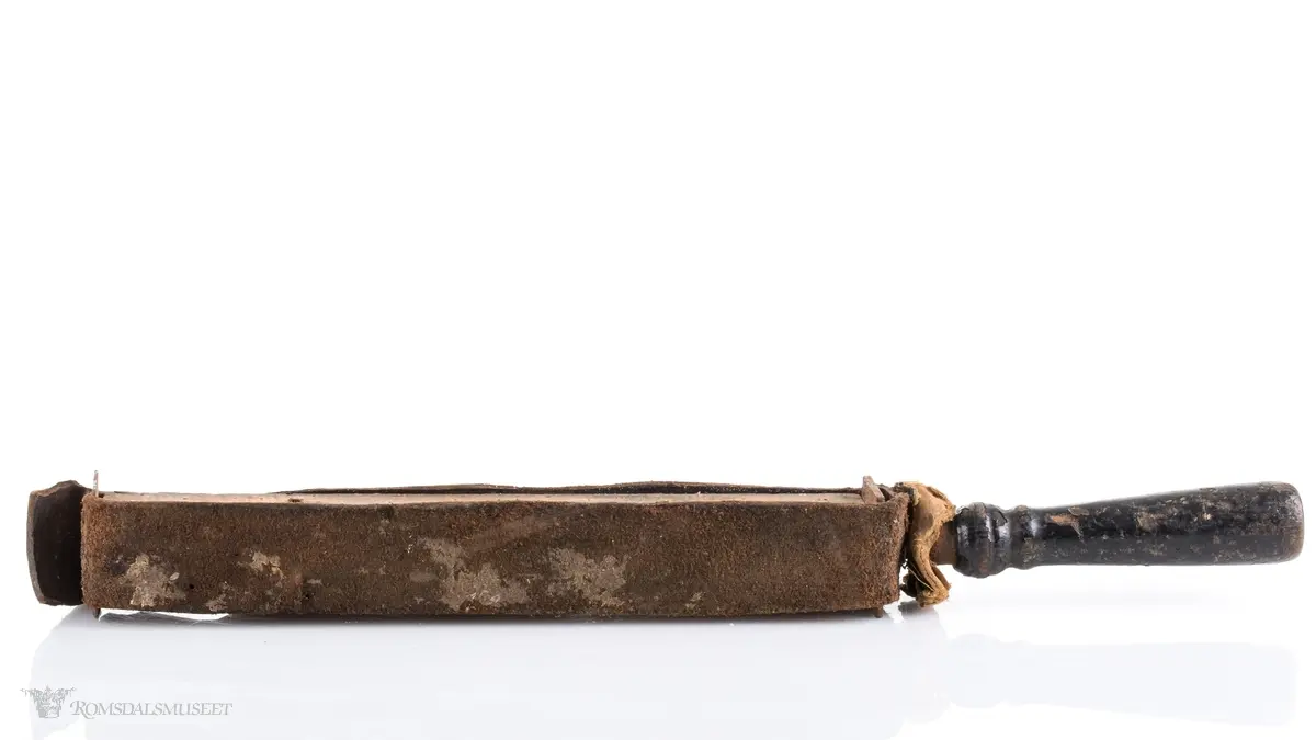 Rektangulær med dreid trehåndtak. Lærrem som går rund hele. Avstivet med metallbeslag foran og bak. Brukt til å stroppe/kvesse barberknivblad før bruk.