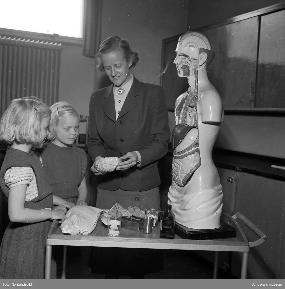 En sjuksköterska förevisar en pedagogisk anatomidocka för två små flickor (tvillingar?) på sjuksköterskeskolan vid Sundsvalls lasarett.