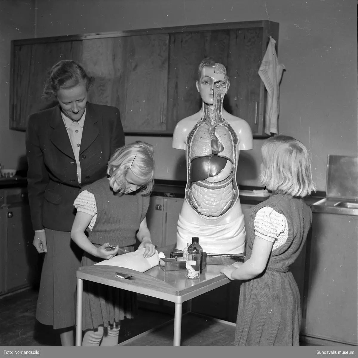 En sjuksköterska förevisar en pedagogisk anatomidocka för två små flickor (tvillingar?) på sjuksköterskeskolan vid Sundsvalls lasarett.