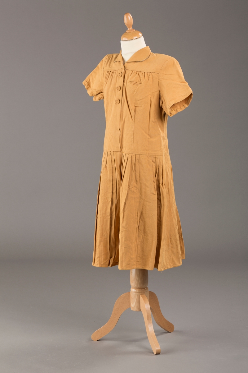 Form: Korte ermer, livet rynket til bærestykke foran, plissert skjørt, tre pynteknapper, en trykknapp, forlomme.