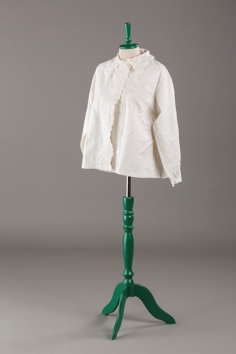 Form: Hoftelang trøye, Lange ermer, krage. Kragen og åpningen foran er kantet med  maskinprodusert blonde. Tre biser på hver side av åpn. foran. Lukkes med to løse knapper.